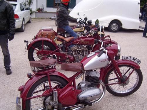 A nostalgic gathering of classic motorcycles, including a maroon Indian and a red-and-silver model, parked on a gravel lot near cars and a van, evokes a vintage enthusiast meetup ideal for motorcycle lovers exploring retro rides.
