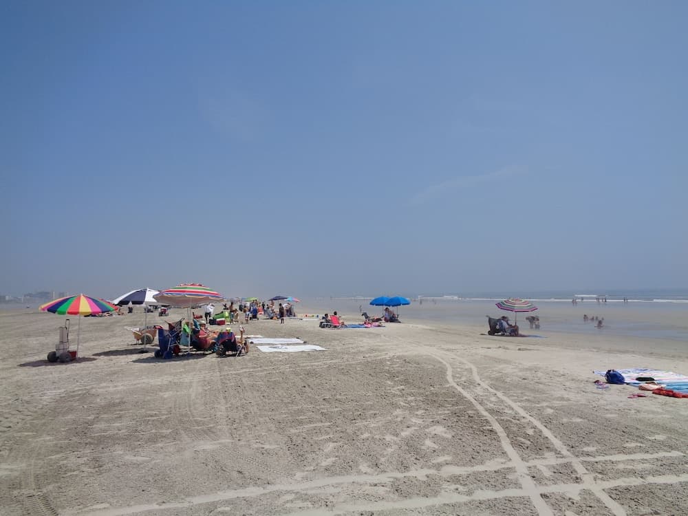 A serene beach scene unfolds under a clear blue sky, featuring colorful umbrellas, scattered beachgoers, and tire tracks in the sand, ideal for a relaxing summer getaway.