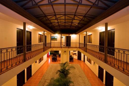 A warmly lit hotel atrium features terracotta floors, a central palm plant, and balconies with black railings, creating an inviting atmosphere ideal for travelers seeking cozy accommodations.