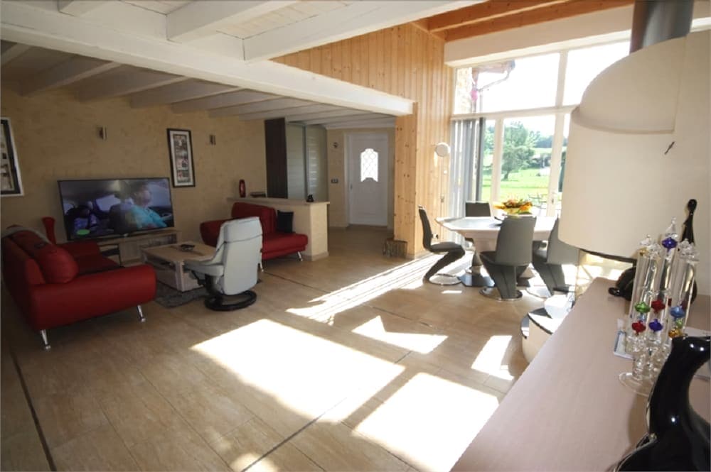 A cozy, sunlit living room with red sofas, a modern dining area, and large windows overlooking greenery, ideal for a relaxing family vacation retreat.