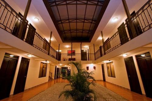 A warmly lit hotel atrium features a central palm plant, wooden floors, and balconies, creating an inviting atmosphere ideal for travelers seeking cozy accommodations.