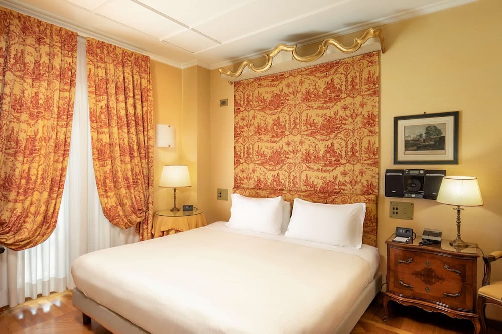 A cozy hotel room features warm yellow walls adorned with intricate red-patterned wallpaper, plush bedding, vintage wooden furniture, and ambient lighting, ideal for travelers seeking comfort and classic charm.