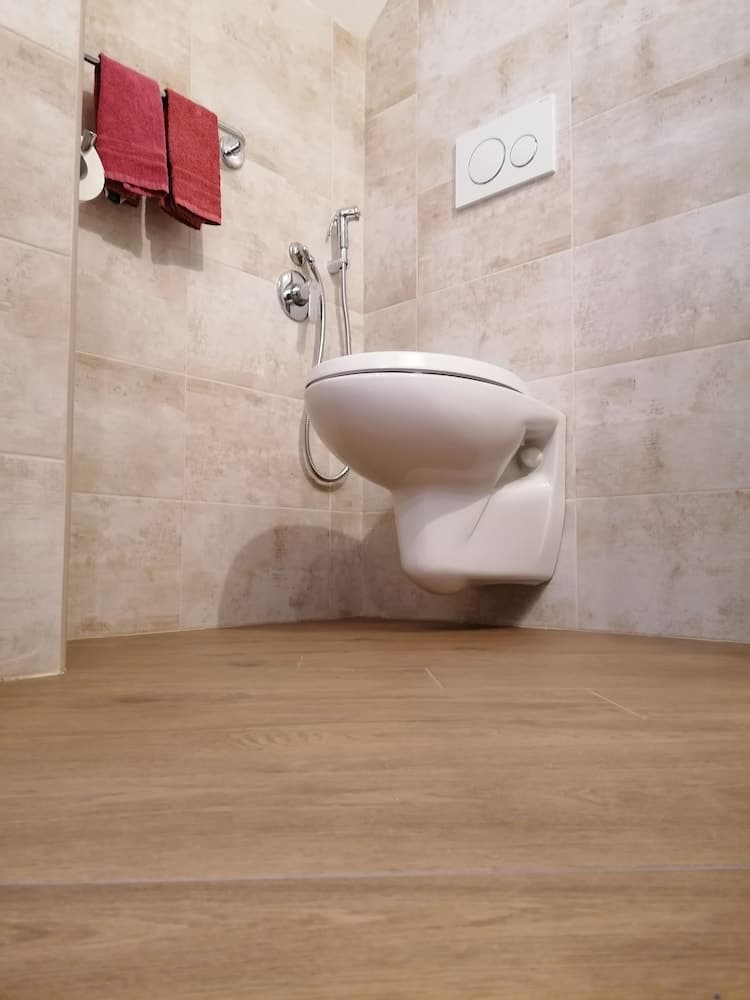 A modern bathroom with beige tiled walls, a wall-mounted toilet, wooden flooring, and red towels creates a clean and cozy atmosphere, ideal for travelers seeking comfort in accommodations.