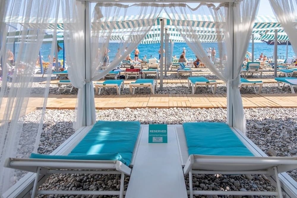 A serene beachfront scene features reserved lounge chairs under sheer curtains on a pebble beach, with turquoise cushions and striped umbrellas enhancing the tranquil seaside atmosphere, perfect for a relaxing vacation getaway.