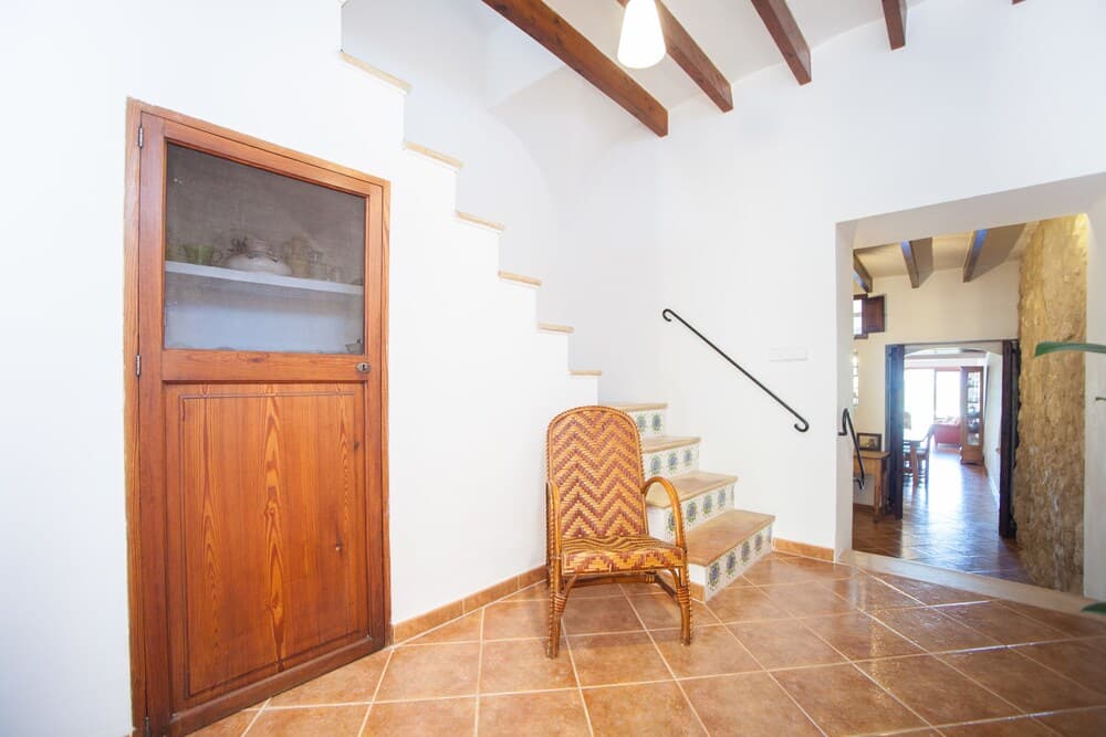 A cozy Mediterranean-style interior features tiled floors, wooden beams, a patterned chair, and a staircase leading to an inviting upper level, perfect for travelers seeking rustic charm in their accommodations.