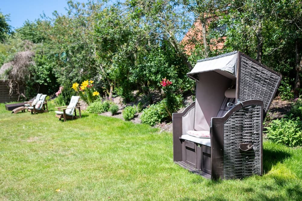 A serene garden scene features a cozy wicker beach chair with a canopy, surrounded by lush greenery, vibrant flowers, and lounge chairs, ideal for a relaxing outdoor retreat.