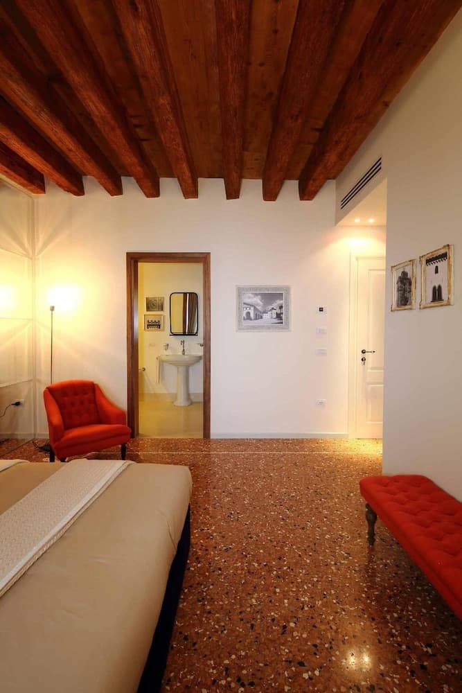 A cozy hotel room features a rustic wooden ceiling, terrazzo flooring, vibrant red seating, and an en-suite bathroom, ideal for travelers seeking charm and comfort.