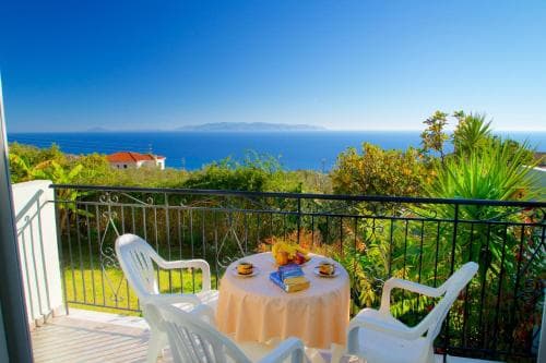 A serene coastal balcony offers breathtaking ocean views, perfect for a tranquil getaway, featuring white chairs, a table set for breakfast, lush greenery, and a distant red-roofed house under a clear blue sky.