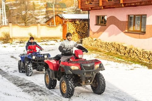 Two individuals enjoy a winter adventure on ATVs in a snowy village setting, surrounded by rustic houses and stacked firewood, ideal for travelers seeking outdoor activities in a serene, cold-weather destination.