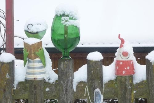 A whimsical snow-covered garden fence adorned with quirky bottle decorations creates a charming winter scene perfect for seasonal photography and festive travel inspiration.