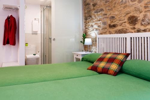 A cozy bedroom with a green bedspread, plaid pillow, stone wall, and en-suite bathroom offers a charming and rustic retreat for travelers seeking comfort in a quaint setting.