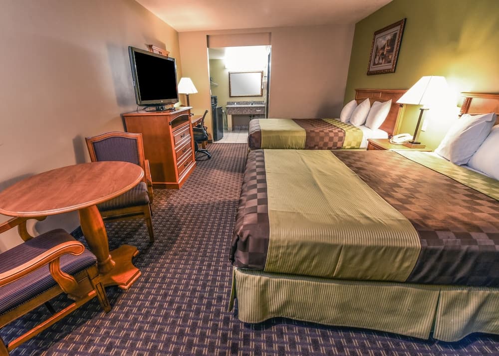 A cozy hotel room features two neatly made beds with green and brown bedding, a wooden table with chairs, a TV on a dresser, and a bathroom in the background, ideal for travelers seeking comfort and convenience.