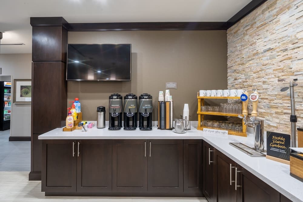 A cozy hotel coffee bar features dark wood cabinets, a stone wall backdrop, and an array of coffee dispensers, creating a welcoming atmosphere for travelers seeking a fresh brew and baked goods.