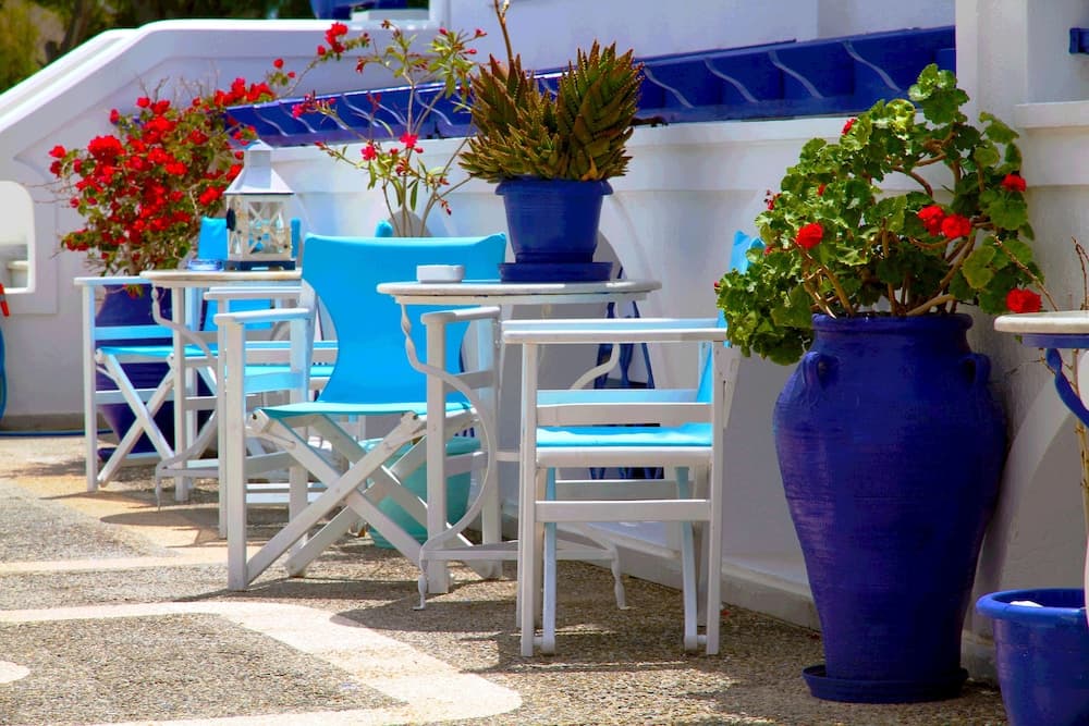 A charming Mediterranean café scene features white tables and blue chairs adorned with vibrant red flowers in large blue pots, evoking a serene and inviting atmosphere perfect for travelers seeking a relaxing seaside retreat.