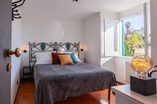 A cozy bedroom with a stylish metal bedframe, vibrant pillows, and a sunny view through large windows, perfect for a relaxing getaway in a modern urban retreat.