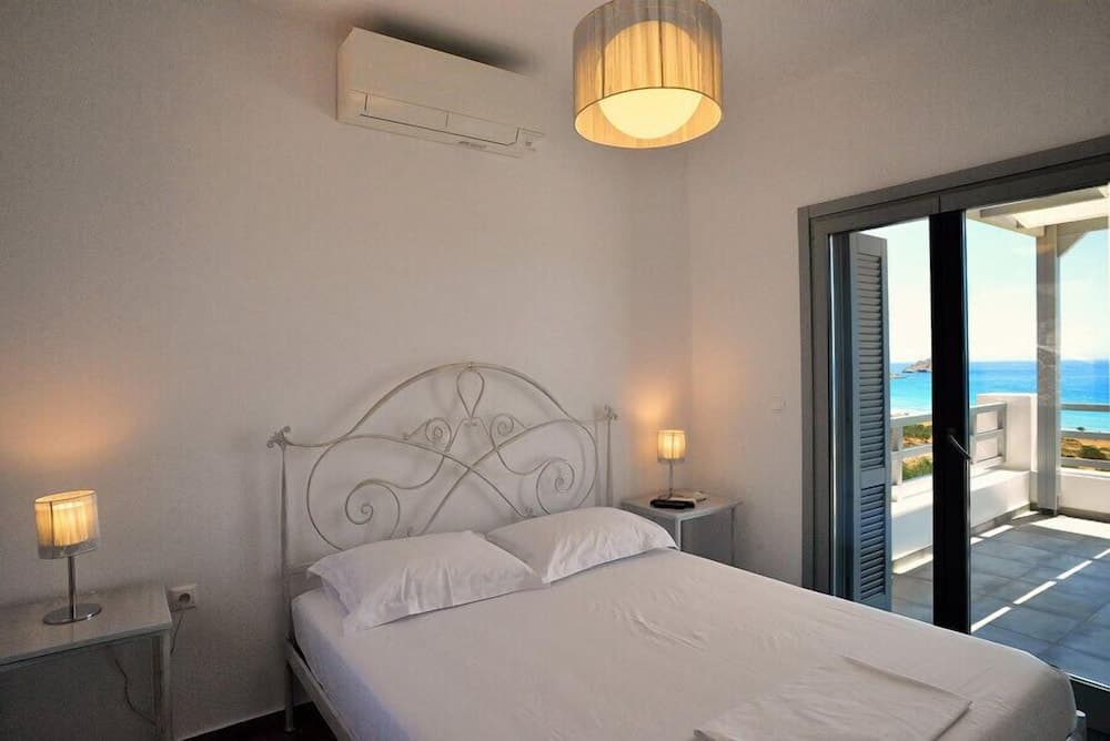 A serene coastal bedroom features a white metal bed, soft lighting, and sliding glass doors leading to a balcony with ocean views, ideal for a relaxing seaside getaway.