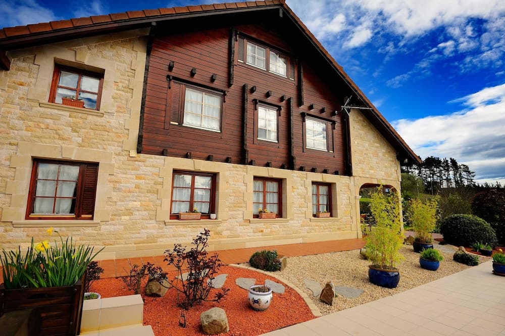 A charming rustic house with stone and wooden paneling, surrounded by a well-maintained garden with vibrant plants and gravel pathways, offers a serene retreat under a picturesque blue sky, ideal for travelers seeking a cozy countryside escape.