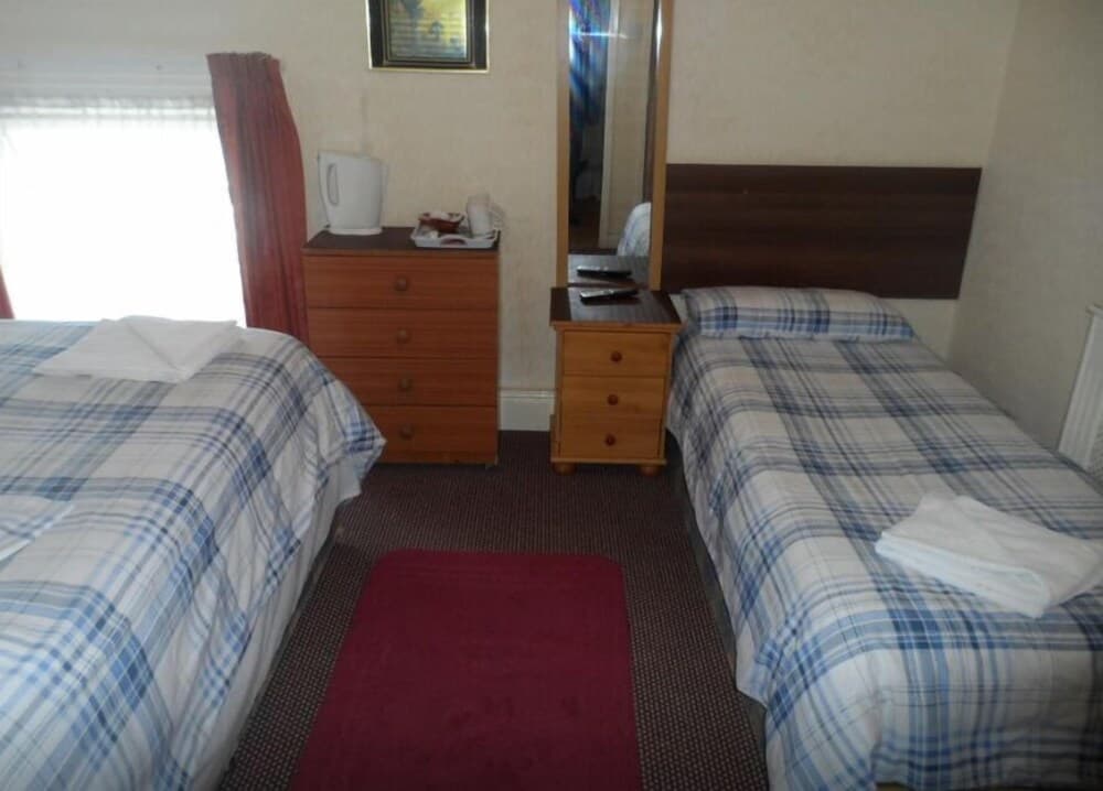 A cozy budget-friendly hotel room features two single beds with blue plaid bedding, wooden furniture, a red rug, and natural light from a window, ideal for travelers seeking simple accommodations.