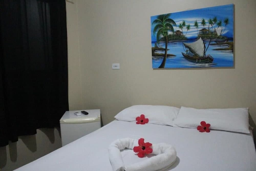 A cozy hotel room features a neatly made bed adorned with red flower decorations and a tropical island painting, creating a serene and inviting atmosphere ideal for travelers seeking relaxation.