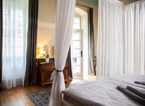 A cozy bedroom with wooden floors, draped white curtains, and a vintage desk by the window invites travelers seeking a serene and elegant retreat.