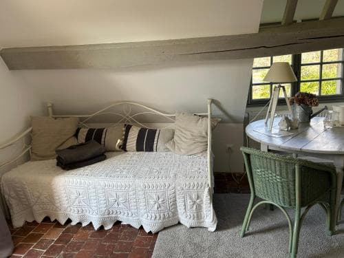 A cozy, rustic bedroom with a white metal bed adorned with patterned bedding, a small round table with a lamp and vase by the window, and exposed wooden beams, ideal for travelers seeking a charming countryside retreat.