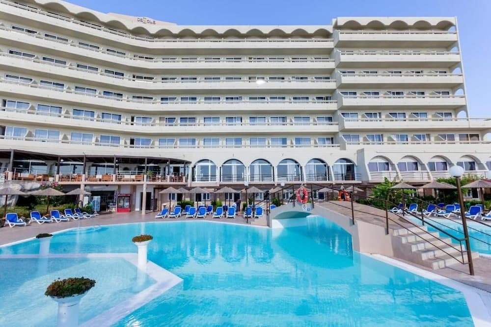 A serene resort scene features a large, curved hotel building overlooking a vibrant blue pool surrounded by lounge chairs and umbrellas, ideal for vacationers seeking relaxation and leisure.