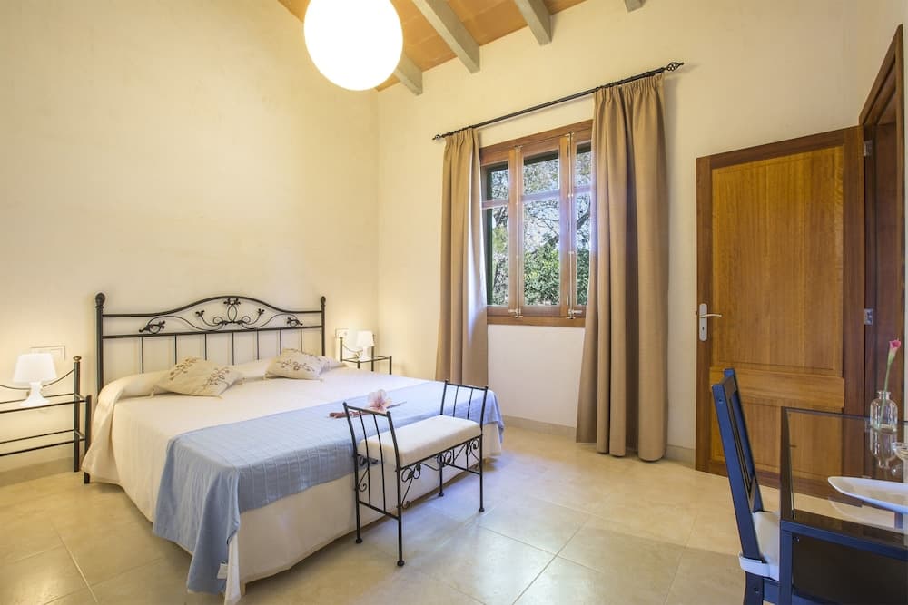A cozy bedroom with a neatly made bed, wooden accents, and a window view of greenery offers a serene retreat for travelers seeking relaxation in a rustic villa setting.