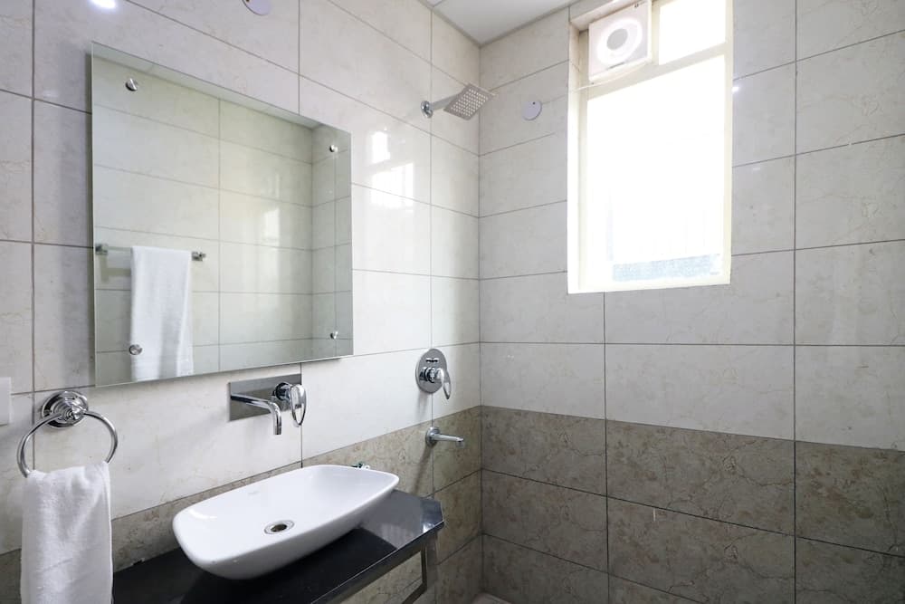 A serene hotel bathroom features sleek white marble tiles, a modern sink, a wall-mounted shower, and a bright window, ideal for travelers seeking comfort and cleanliness.