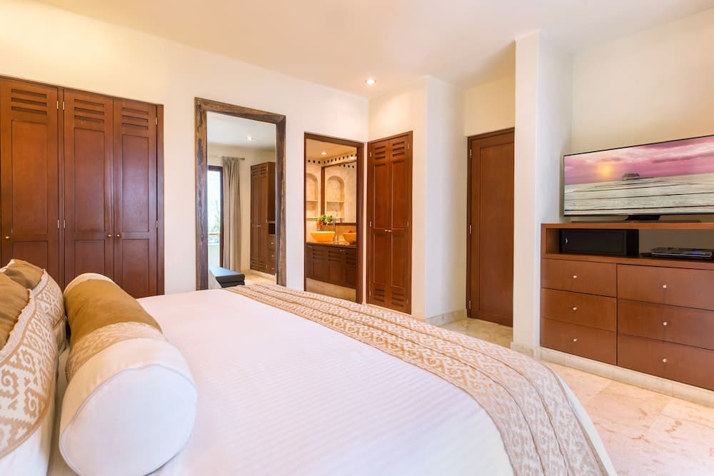 A cozy hotel bedroom features a neatly made bed, wooden furnishings, a mounted TV displaying a serene sunset, and an open doorway leading to a bathroom, ideal for travelers seeking comfort and relaxation.