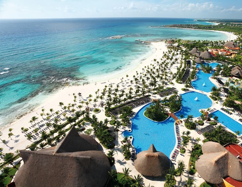 An aerial view of a luxurious all-inclusive resort along the pristine beaches of the Caribbean, featuring expansive pools, thatched-roof structures, and lush palm trees, ideal for a tropical getaway in Playa del Carmen, Mexico.