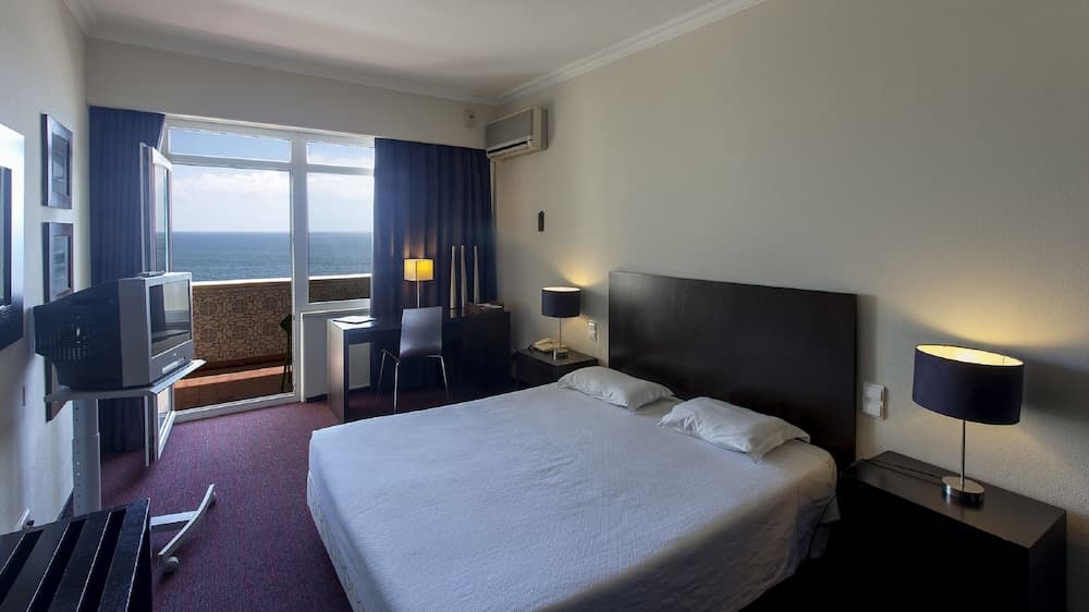 A cozy hotel room with a serene ocean view through sliding glass doors, featuring a neatly made bed, warm lighting from bedside lamps, and a television, ideal for travelers seeking relaxation by the sea.