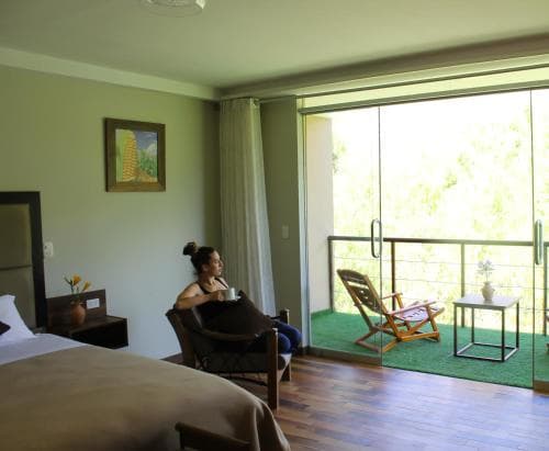 A serene hotel room with a cozy ambiance features a woman enjoying coffee by a large window overlooking lush greenery, complemented by comfortable seating and a small balcony, ideal for travelers seeking relaxation and nature proximity.
