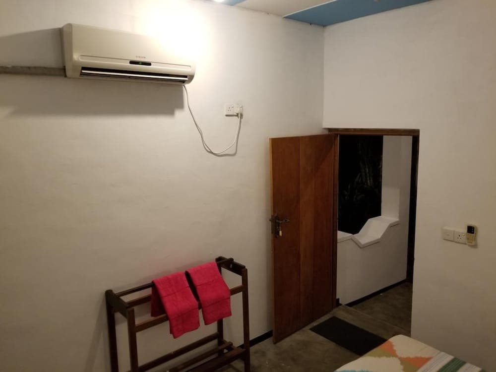 A cozy hotel room corner features a wooden door leading to a bathroom, a towel rack with red towels, an air conditioner, and a table with a colorful cloth, ideal for travelers seeking comfort and convenience.