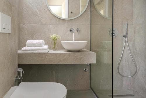 A serene hotel bathroom features a modern sink with neatly folded towels, a vase of flowers, a glass-enclosed shower, and round mirrors, ideal for travelers seeking comfort and style.