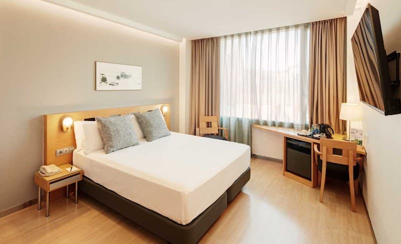 A cozy hotel room in Barcelona's Eixample district features a neatly made bed, wooden furnishings, and soft natural light filtering through sheer curtains, ideal for travelers seeking comfort and convenience.