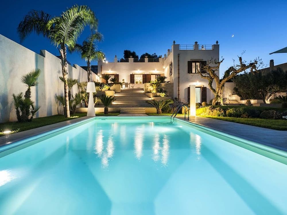 A serene evening at a luxurious villa features a glowing pool, palm trees, and a moonlit sky, ideal for a tranquil getaway.