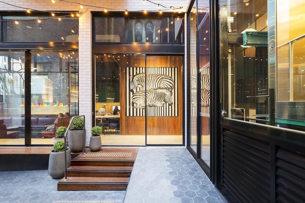 A modern urban café with hexagonal tiled floors, string lights, and a striking zebra-patterned glass wall invites travelers to relax in its stylish and cozy atmosphere.