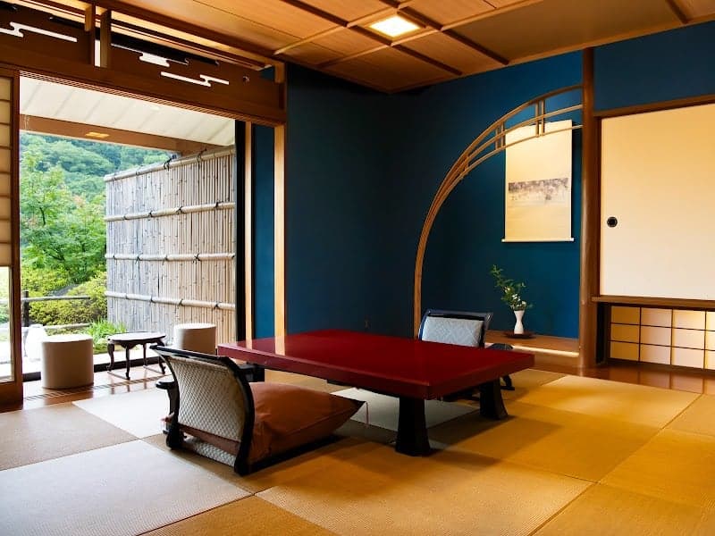 A serene Japanese ryokan room features tatami flooring, a low red table with cushioned chairs, and sliding doors opening to lush greenery, ideal for travelers seeking traditional relaxation and cultural immersion.