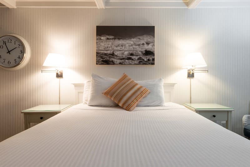 A cozy hotel room in Cannon Beach features a neatly made bed with white linens, a striped pillow, wall-mounted lamps, and a serene black-and-white ocean artwork, ideal for travelers seeking relaxation near the Oregon coast.