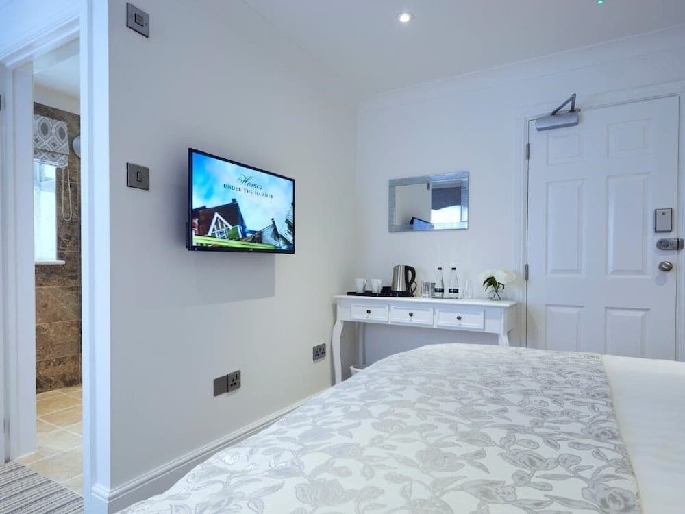 A cozy hotel room features a neatly made bed, a mounted TV displaying a serene scene, a small desk with refreshments, and an adjacent bathroom, ideal for travelers seeking comfort and convenience.