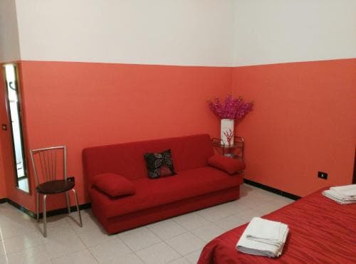A cozy hotel room features a vibrant red sofa against an orange-painted wall, complemented by a simple chair, a vase of purple flowers, and neatly folded towels on the bed, ideal for travelers seeking comfort in a minimalist setting.
