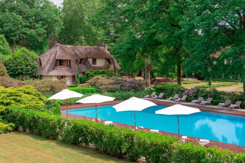 A serene outdoor pool surrounded by lush greenery and thatched-roof cottages offers a tranquil retreat, perfect for relaxation and leisure travel.