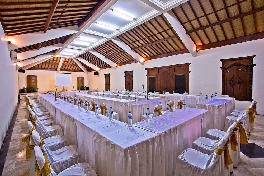 A spacious conference room with wooden-beamed ceilings, neatly arranged tables draped in white linens, and chairs adorned with gold ribbons, creates an elegant setting ideal for professional events and meetings.