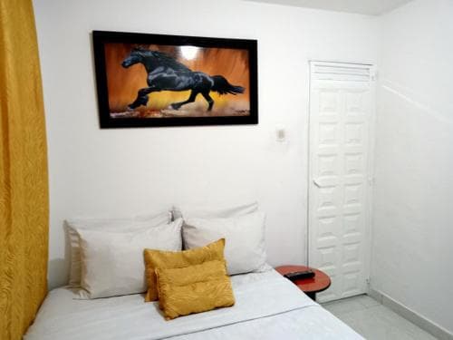 A cozy bedroom with a vibrant horse painting above the bed, white walls, a yellow curtain, and a small round table creates a serene atmosphere ideal for travelers seeking a peaceful retreat.