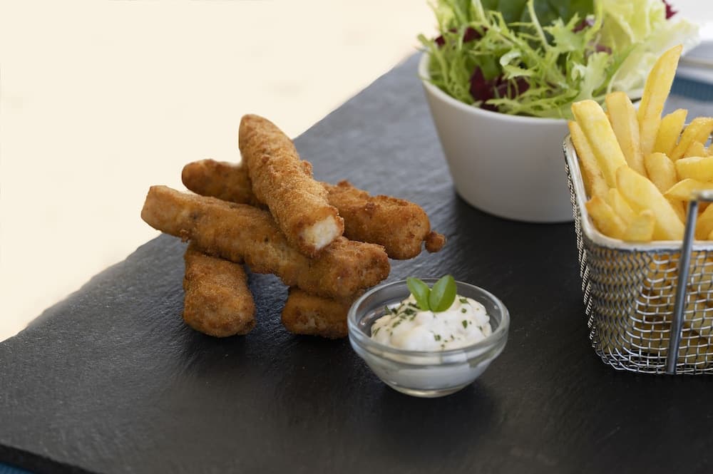 A slate platter showcases crispy fish sticks paired with golden fries in a wire basket, a fresh salad bowl, and creamy dipping sauce garnished with herbs, ideal for a casual dining or beachside snack experience.