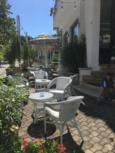 A cozy outdoor café with white wicker furniture, lush greenery, and a thatched umbrella invites travelers to relax under the sunny sky, perfect for a leisurely coffee break or a quiet afternoon read.