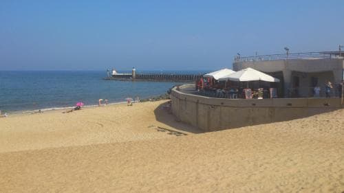 A sunny beach scene features a sandy shore with scattered beachgoers, a pier extending into calm blue waters, and a shaded seating area under white umbrellas near a concrete structure, ideal for a relaxing seaside getaway.