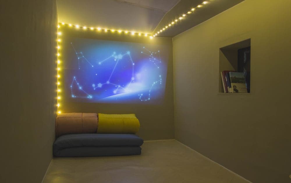 A cozy indoor relaxation space features a starry constellation projection on the wall, surrounded by warm string lights, with a comfortable cushioned seating area and a bookshelf, ideal for travelers seeking a serene and immersive experience.