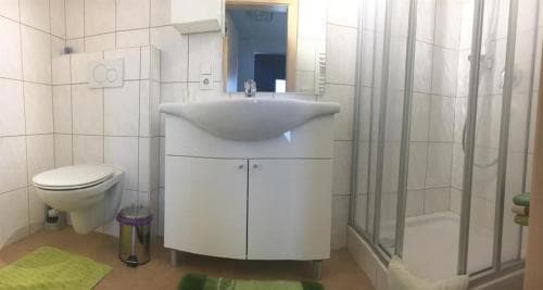 A clean, compact bathroom with white tiled walls, a modern sink cabinet, a toilet, a glass-enclosed shower, and green accents creates a functional and inviting space ideal for travelers seeking comfort in accommodations.