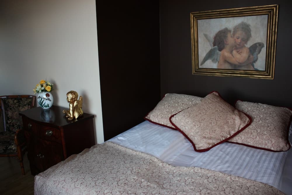 A cozy bedroom features a neatly made bed adorned with patterned pillows, a dark wooden nightstand topped with a vase of flowers and a golden figurine, and a framed angel painting on the wall, ideal for travelers seeking a serene and artistic retreat.